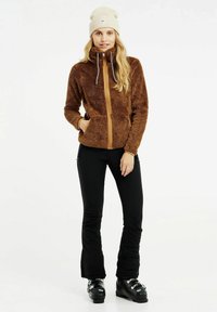 Bruine, fuzzy jas met een beige rits, hoge kraag en twee voorkeurszakken; gecombineerd met zwarte broek en zwarte laarzen. Beigekleurige beanie inbegrepen.