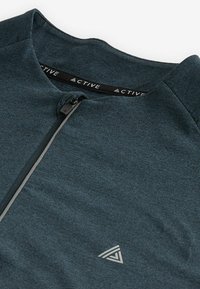 Dunkeltürkises Sportshirt mit hohem Kragen, kurzem Reißverschluss und dezentem Logo am Nacken. Weicher, strukturiert Stoff.