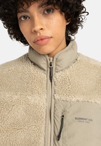 Beige fleecejacka med strukturerad krämfärgad panel, hel dragkedja och sidofickor. Har svart dragkedja och tryckt logo på vänster bröst.