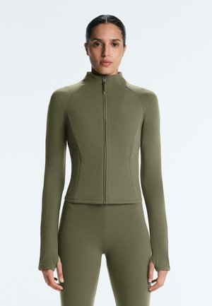 Žena oblečená v priliehavom olivovozelenom zipsovom obere a zodpovedajúcich legínach, stojaca pred jednoduchým svetlým pozadím.