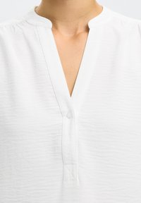 White textured shirt with a V-neckline and a short stand collar, featuring a single visible button on the front placket.