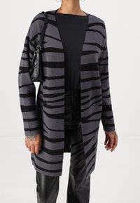 Szary kardigan z czarnymi tygrysimi paskami, wykonany z miękkiego materiału. Otwarty przód, w połączeniu z czarnym topem i skórzanymi spodniami. Marszczona faktura.