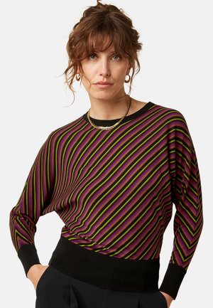 Vrouw met bruin haar in een gestreepte paarse, groene en zwarte trui en zwarte broek, met gouden oorringen en een kettingketting.