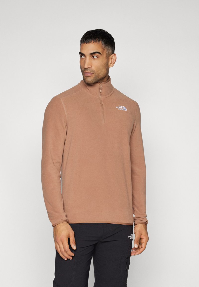 Brauner Fleece-Pullover mit halbem Reißverschluss, ausgestattet mit einem hohen Kragen und kleinem Logo auf der Brust. Das Modell trägt dunkle Hosen dazu.