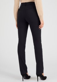 Pantalones negros ajustados de corte slim, con textura suave, cintura alta y con dos bolsillos traseros. Combinados con zapatos de tacón alto negros.