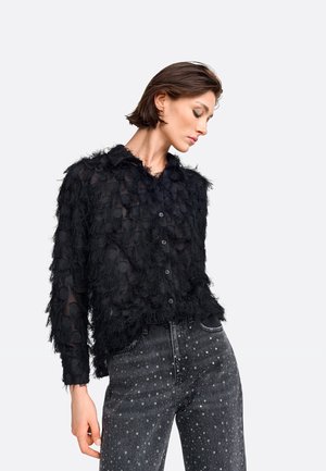 Schwarze Bluse mit Federstruktur, Kragen, Knöpfen auf der Vorderseite und transparenter Gestaltung, kombiniert mit dunklen Jeans mit silbernen Verzierungen.