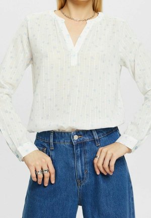 Witte blouse met lange mouwen en een subtiel blauw bloemenpatroon, v-hals en gestreepte textuur. Gecombineerd met hoge blauwe spijkerbroek.