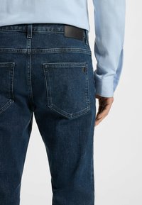 Mørkeblå denimjeans med en teksturert overflate, har to baklommer, lærmerke ved livlinjen og en rett bendesign.