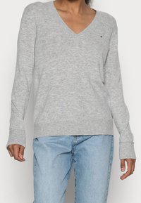 Grauer V-Ausschnitt-Pullover aus weichem Strickstoff, mit gerippten Bündchen und Saum. Enthält ein kleines Logo-Detail auf der Brust.