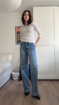 Nuori nainen seisoo sisätiloissa valkoisessa pitkähihaisessa paidassa, sinisissä farkuissa ja mustissa teräväkärkisissä kengissä huoneessa, jossa on sänky ja vaatekaappi.