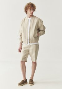 Beige bomberjakke i let stof, hvid t-shirt, matchende shorts og brune slip-on sko. Afslappet pasform med ribbet fald og kant.