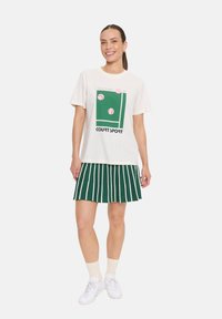 T-shirt blanc avec un graphisme de court de tennis vert et des balles roses. Associé à une jupe plissée verte avec des rayures blanches verticales. Baskets blanches.