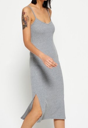 Kvinde i ærmeløs, grå og hvid stribet midi-kjole med sideslids, der viser tatoveret overarm, gående mod en ensfarvet baggrund.