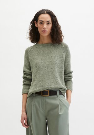 Femme aux cheveux bouclés et foncés portant un pull en maille vert olive et un pantalon large assorti avec une ceinture noire, debout avec une main dans la poche.