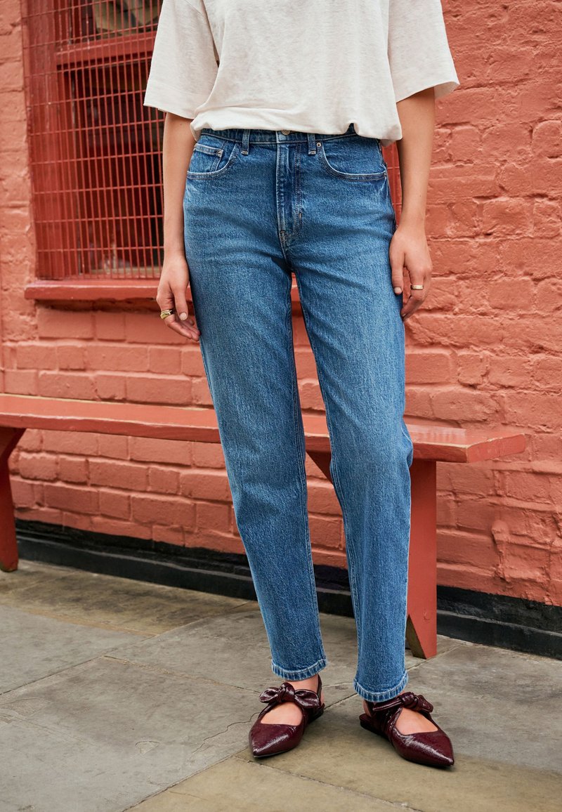 Persona valkā augsta jostasvietas zilas džinsas, baltu īsroku topu un tumši sarkanas adātas kurpes ar smailu degunu, stāv pie sarkanā ķieģeļu mūra un sola.