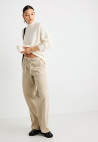 Kreminės spalvos golfas iš megztinio, laisvo kirpimo, derinamas su smėlio spalvos aukštos liemens kelnėmis su reguliuojamomis stringais ir tiesiomis kojomis, dėvimas su juodais batais.