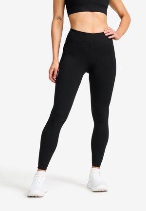 Kvinna klädd i svarta högmidjade leggings och vita sneakers, stående med en hand på höften och synlig arm-tatuering mot vit bakgrund.