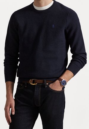 Mand iført en mørk marineblå striktrøje over en hvid undertrøje, mørke jeans, brun læderbælte og et armbåndsur med blå urskive og brun rem.