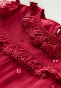 Tessuto rosso con bordo in pizzo floreale ricamato e piccoli accenti di paillettes rosa, che mette in mostra la texture e la cucitura dettagliata del tessuto.