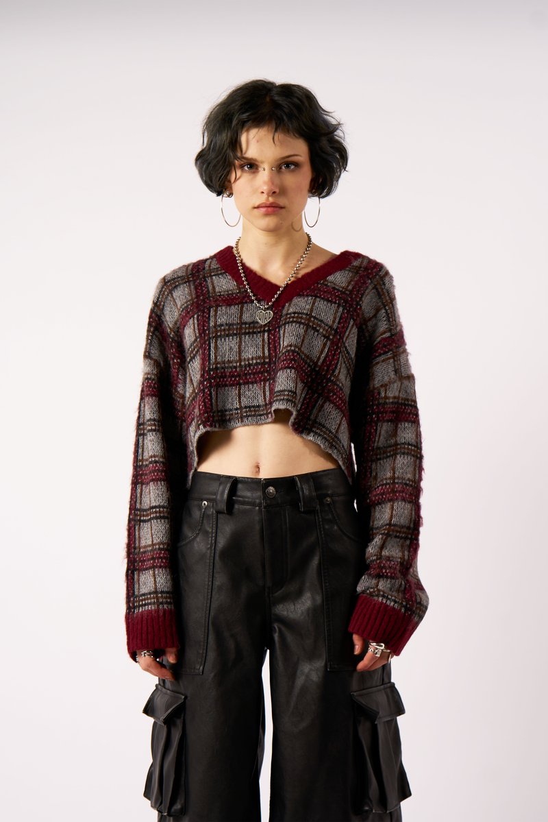 Ung person med kort mørkt hår iført en cropped ternet sweater og sorte cargo bukser, står op ad en ensfarvet lys baggrund.