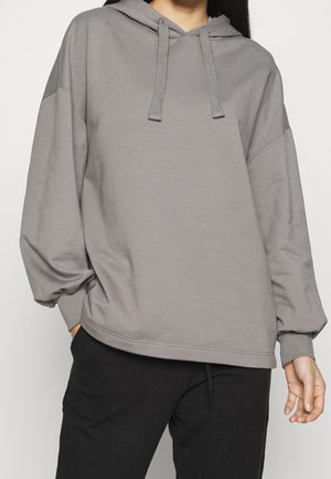 Persoon draagt een losse grijze hoodie met trekkoorden en zwarte broek, staand tegen een effen lichte achtergrond.