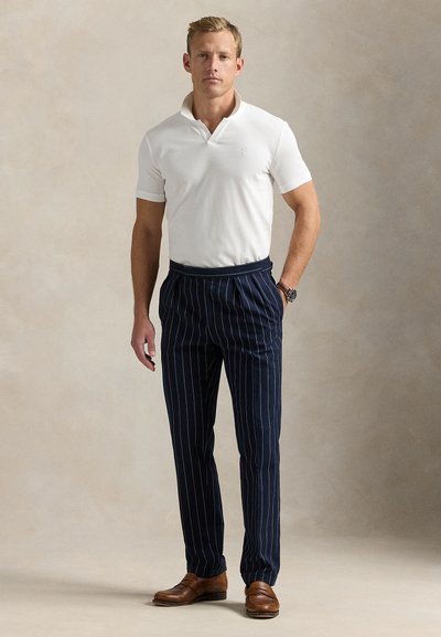 Balti polo marškinėliai, tamsiai mėlyni dryžuoti kelnės, rudi odiniai batai. Paprastas dizainas su klasikiniu kirpimu ir subtiliomis tekstūromis.