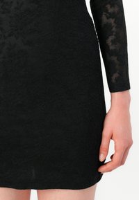Robe en dentelle noire avec des manches longues, un motif floral texturé et une silhouette ajustée qui s'arrête au-dessus du genou.