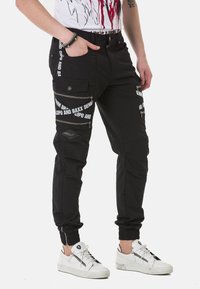 Schwarze Cargo-Hosen mit mehreren Taschen, Reißverschlüssen und weißen Textmustern. Eng an den Knöcheln mit einem strukturierten Stoff. Kombiniert mit weißen Sneakern.