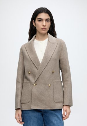 Vrouw met lang donker haar draagt een beige dubbelrijige wollen blazer met gouden knopen, een witte blouse en een blauwe spijkerbroek op een effen lichte achtergrond.