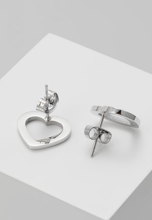 Boucles d'oreilles clous en forme de cœur ouvert ton argent avec attaches papillon, placées sur une surface blanche.