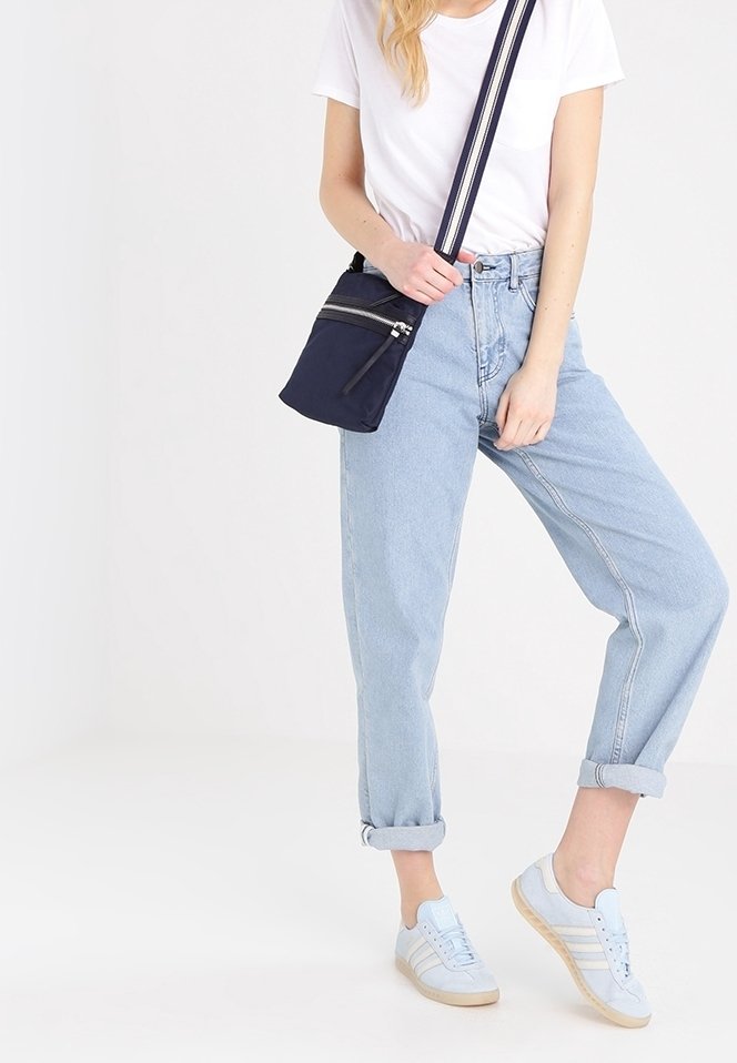 Femme en jean bleu clair retroussé, baskets blanches, t-shirt blanc et sac à bandoulière marine se tenant sur une jambe devant un fond blanc.