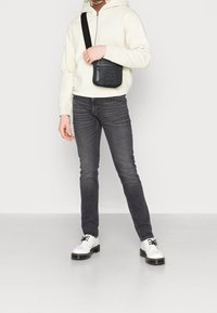 Sac bandoulière noir, sweat à capuche crème, jean gris foncé et chaussures blanches à lacets avec semelles noires. Textures lisses, coupe décontractée.