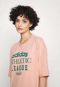 Rožinė medvilninė marškinė su laisvu siluetu, dekoruota didelėmis žaliomis raidėmis "adidas ATHLETIC LEAGUE" ir logotipu. Trumpomis rankovėmis ir apvalia iškirpte.