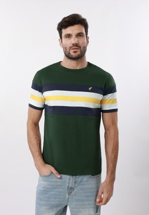 Mężczyzna z brodą w zielonej koszulce z poziomymi paskami w kolorze granatowym, białym i żółtym oraz jasnoniebieskich dżinsach, stojący na białym tle.