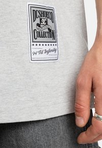 Graues T-Shirt-Material mit einem schwarz-weißen Etikett mit Text. Das Etikett hat eine rechteckige Form, eingerahmt von einem dekorativen Design und Sternen.