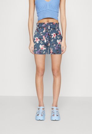 Szczupła sylwetka ubrana w niebieski szydełkowy crop top, kwieciste, krótkie spodenki z wysokim stanem i zawiązanym paskiem oraz masywne niebieskie sandały z odkrytymi palcami, stojąca na jednolitym tle.