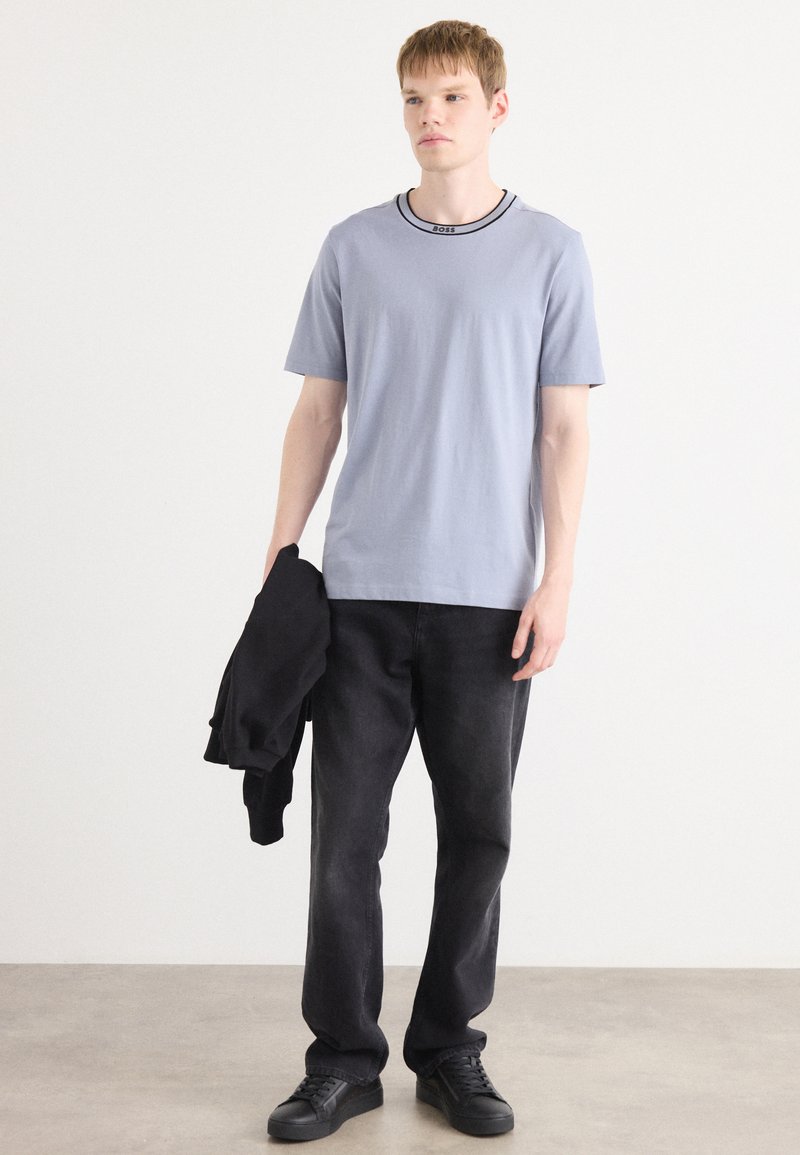 T-shirt azzurro chiaro a maniche corte con rifinitura nera al collo, abbinato a pantaloni neri e sneakers nere. Il modello tiene un maglione nero.