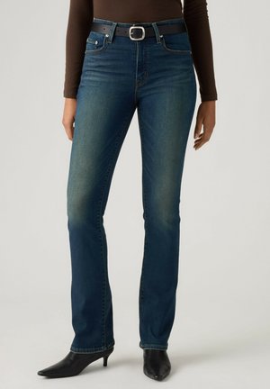Person iført blå, lige ben jeans med sort bælte, brun langærmet top og sorte støvler med hæl, stående mod en ensfarvet baggrund.