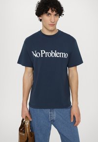 Tyrkysové bavlněné tričko s krátkým rukávem a kulatým výstřihem, s bílým nápisem "No Problemo". Model drží hnědou tašku.