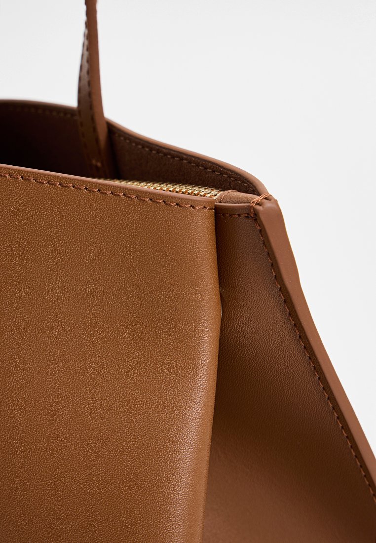 Zbliżenie brązowej skórzanej torby, ukazujące detale zszycia oraz część złotego zamka pod otwartą krawędzią.