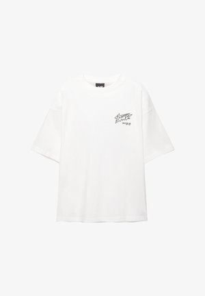 Valkoinen ylisuuri T-paita, joka on valmistettu puuvillasta, lyhyillä hihoilla ja pyöreällä kaula-aukolla. Vasemmassa rinnassa on pieni musta graafinen kuvio.