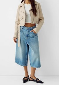 Giacca corta in pelle beige con cerniere argentate, abbinata a pantaloncini in denim azzurro a gamba larga e ballerine nere con punta affilata e borchie.