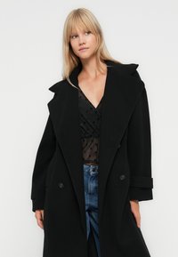 Blondyna w czarnym, oversize'owym płaszczu na przezroczystej czarnej bluzce w kropki i niebieskich dżinsach na tle jednolitego białego tła.