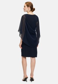 Granatowa sukienka z przezroczystym detalem w kształcie peleryny opadającym z ramion. Zapięcie na plecach z zamkiem błyskawicznym. Gładki materiał i dopasowany krój. Sandały na wysokim obcasie.