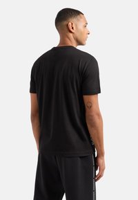 Camiseta negra de manga corta con cuello redondo, textura suave y detalles de costura en contraste. Tatuaje visible en el antebrazo derecho.