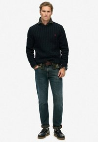 Junger Mann in einem dunklen, strukturierten Pullover über einem grünen Karoshemd, mit blauen Jeans, die an den Knöcheln umgeschlagen sind, und schwarzen Schnürschuhen, der steht.