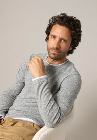 Grauer Pullover mit langen Ärmeln und strukturiertem Finish, mit weißen Akzenten am Ausschnitt. Die Person sitzt vor einem neutralen Hintergrund.