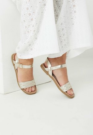 Metallic guld sandaler med et flettet strop, flad sål og justerbar ankelrem, parret med en hvid, broderet kjole.