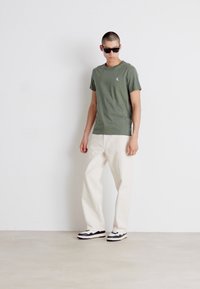 Tricou din bumbac verde măsliniu, cu un mic logo pe piept, asortat cu pantaloni largi de culoare crem și adidași în nuanțe de negru și alb.