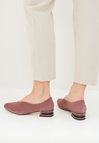 Pantofi din suede, mov, cu toc jos și vârful rotund, având un toc sculptat unic, așezați pe fundalul unor pantaloni deschiși pentru contrast.